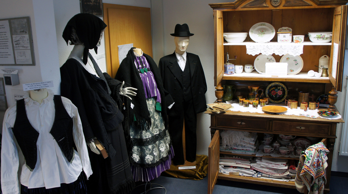 Vier Ausstellungspuppen in Trachtenkleidung und ein Schrank &quot;Wiener Kredenz&quot; geöffnet mit Inhalt, wie Geschirr und Wäsche.